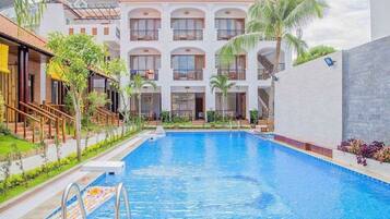 A swimming pool with a clear blue water, surrounded by a white building with balconies and palm trees.