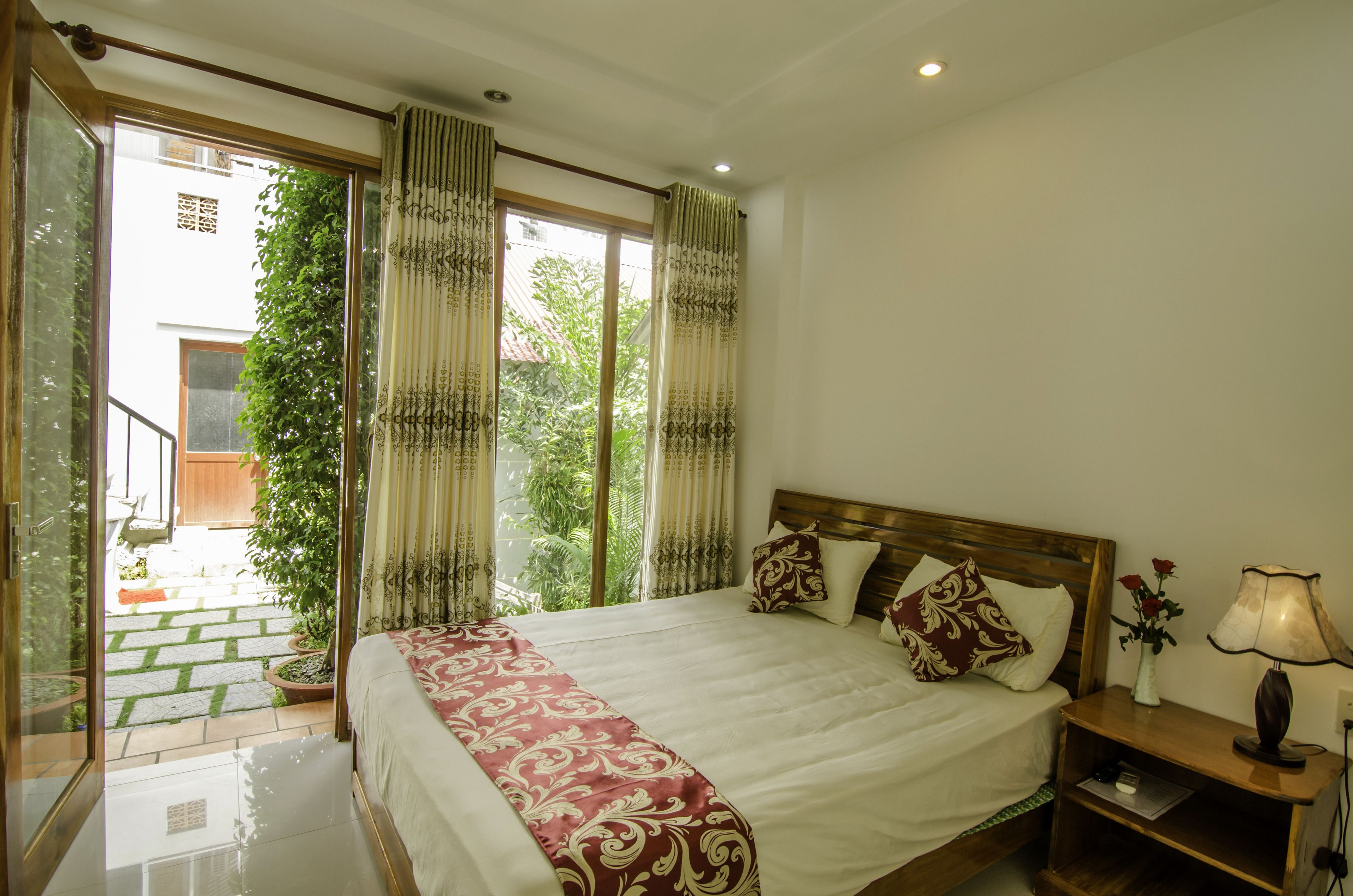 A hotel room with a large bed, a nightstand, a lamp, and a view of a garden through the window.