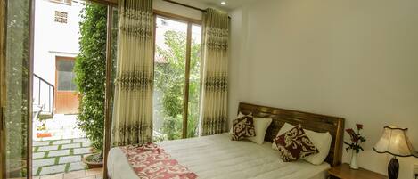 A hotel room with a large bed, a nightstand, a lamp, and a view of a garden through the window.