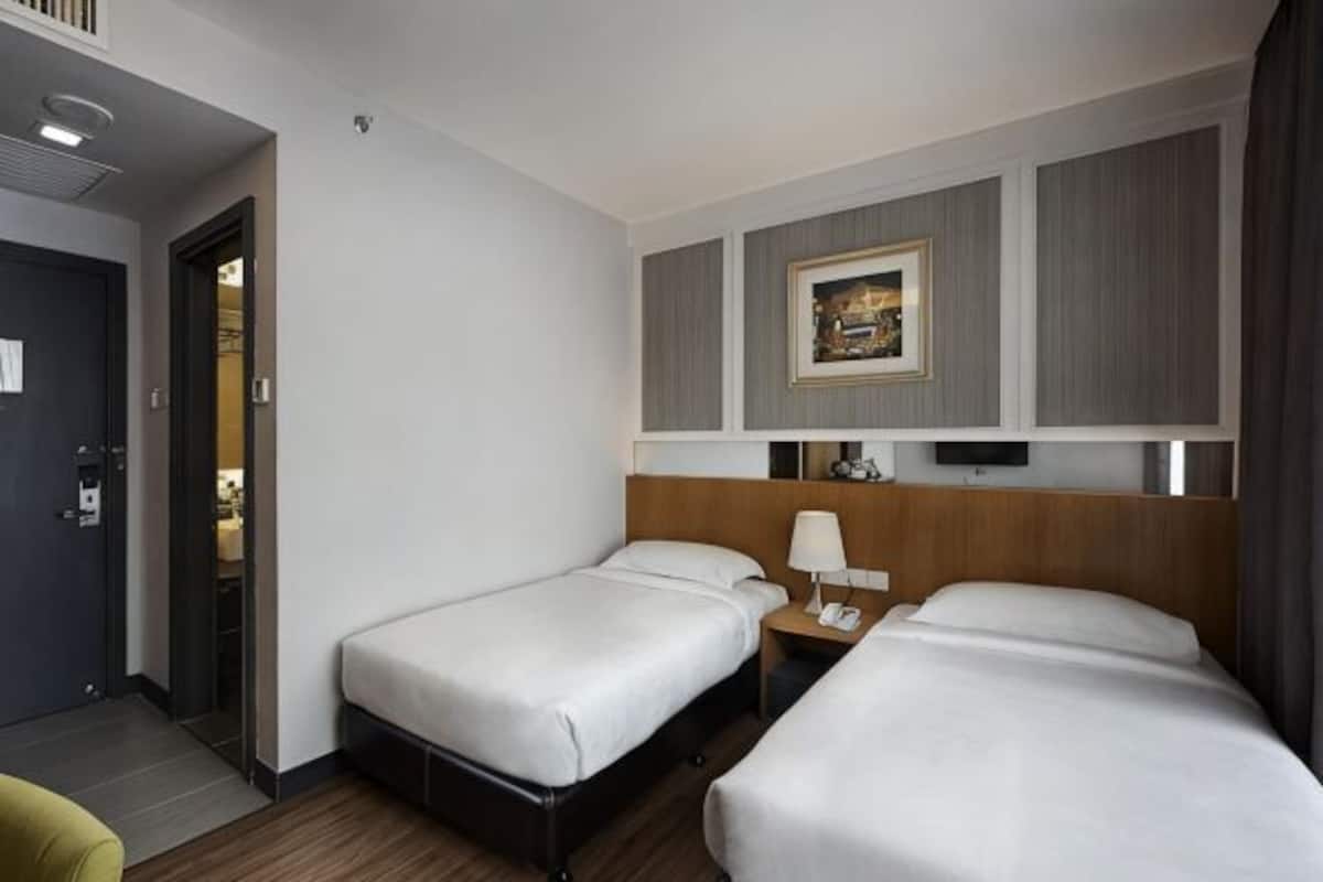 A hotel room with two single beds, a bedside table with a lamp, a wall-mounted TV, and a door leading to another room.