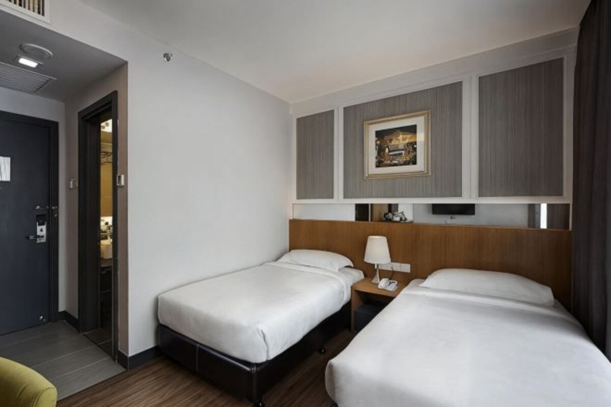A hotel room with two single beds, a bedside table with a lamp, a wall-mounted TV, and a door leading to another room.