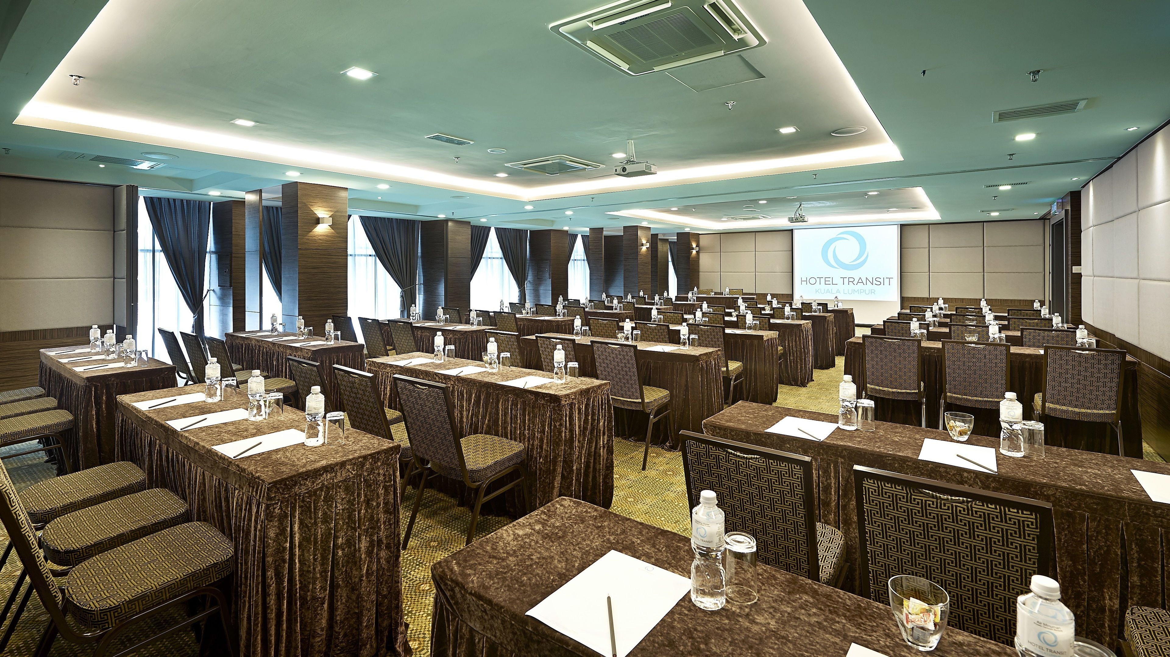 Un salón de conferencias de hotel con varias mesas y sillas, equipado con botellas de agua y blocs de notas, y una pantalla de presentación al fondo.