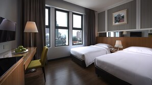 Habitación de hotel con dos camas, un escritorio con televisor, una silla y una ventana con vista a la ciudad.