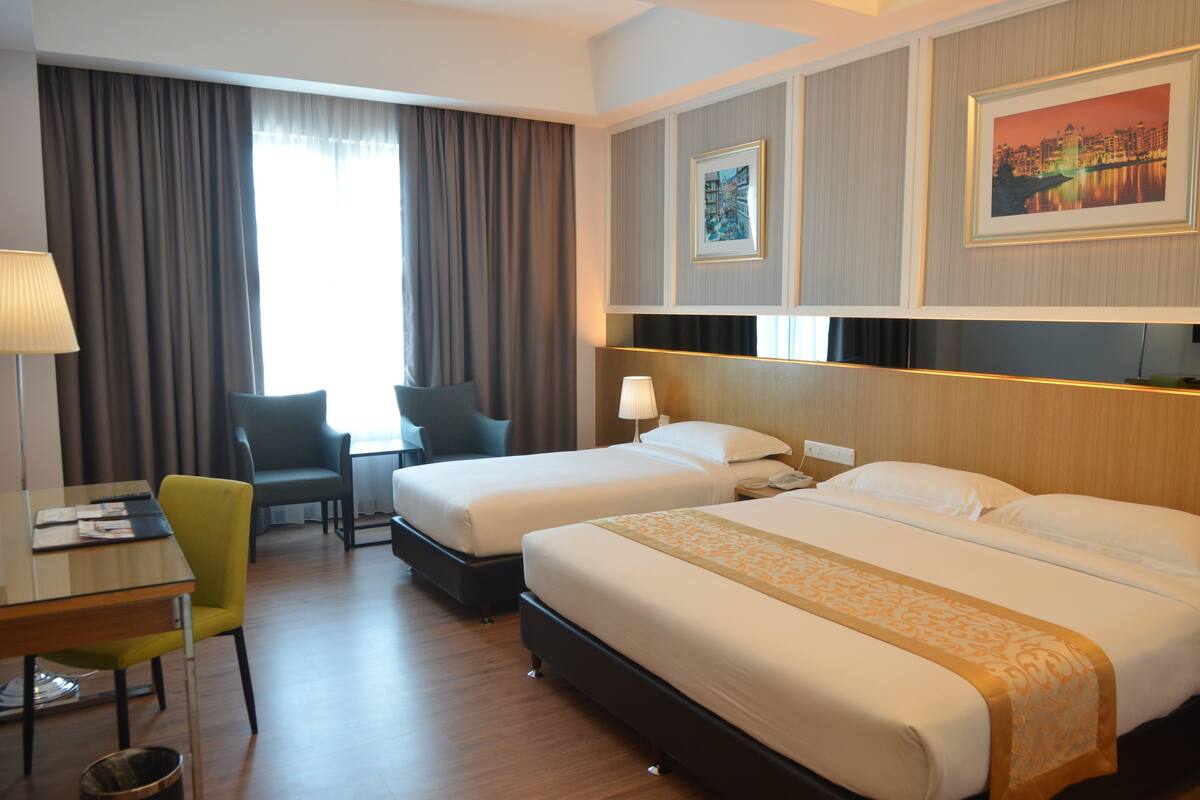 A hotel room with two beds, a desk with a chair, a lamp, and a view of a cityscape through the window.