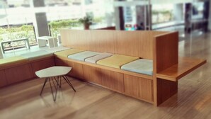 Un banco moderno de madera con asientos acolchados y una pequeña mesa en un espacio luminoso y amplio, con grandes ventanales.