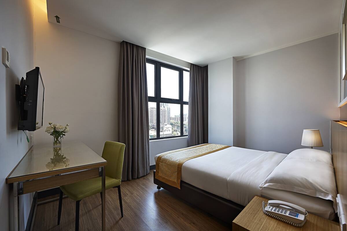 A hotel room with a bed, bedside table with a phone, a desk with a chair, a window with curtains, and a view of buildings outside.
