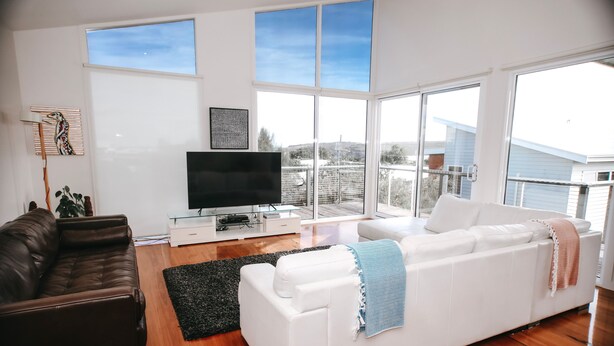 A modern living room with a brown leather sofa, a white sofa with a blue throw, a flat-screen TV, and a large window offering a view of the city. - Anchors Beach House (Port Campbell)
