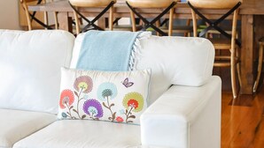 A white leather sofa with a colorful floral pillow, a wooden dining table with chairs, and a kitchen area with dark cabinets and a white stove. - Anchors Beach House (Port Campbell)
