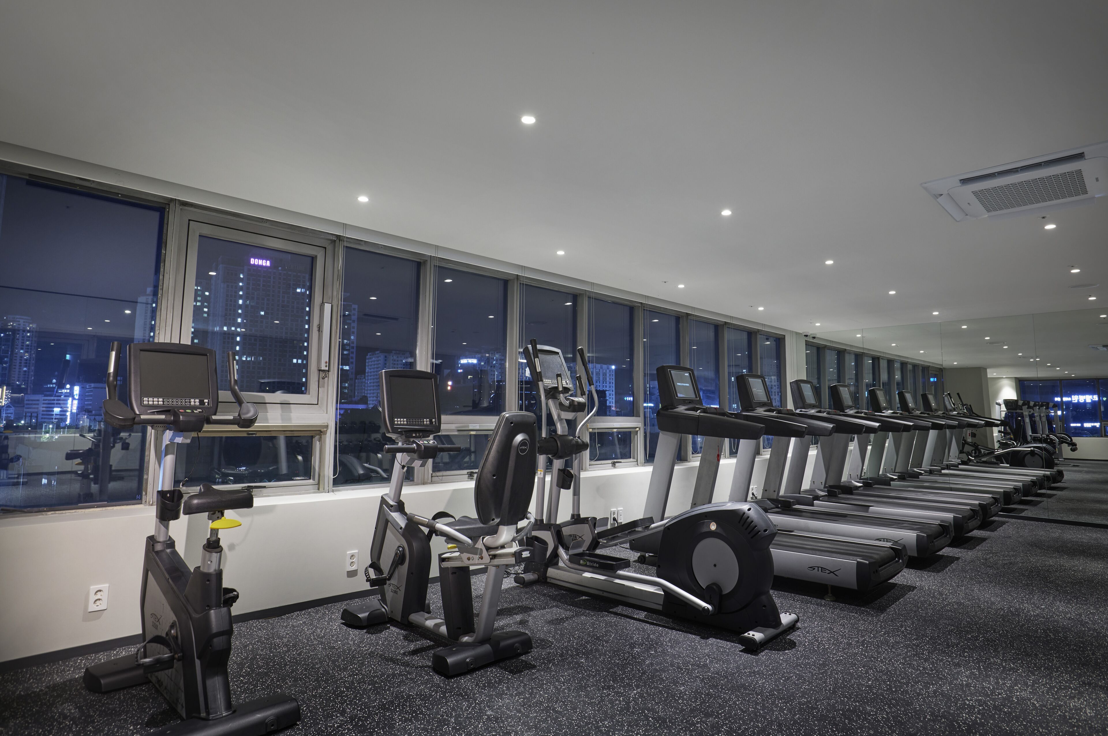Et fitnesscenter med flere løbebånd og træningsmaskiner, store vinduer med udsigt over byen og et tæppe belagt gulv.