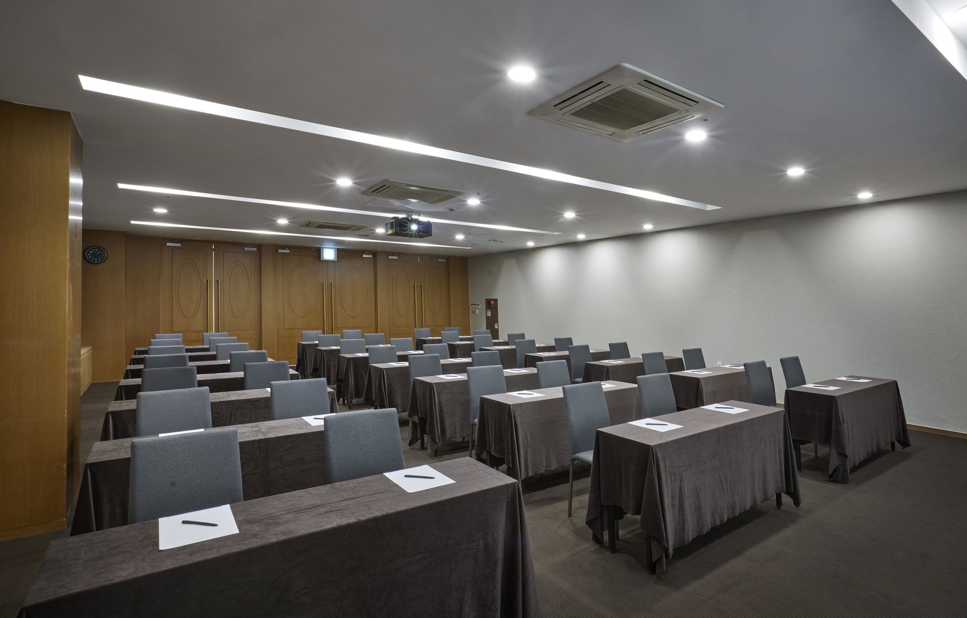 Un salón de conferencias con filas de mesas y sillas, equipado con un proyector e iluminación del techo.
