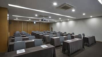 Un salón de conferencias con filas de mesas y sillas, equipado con un proyector e iluminación del techo.