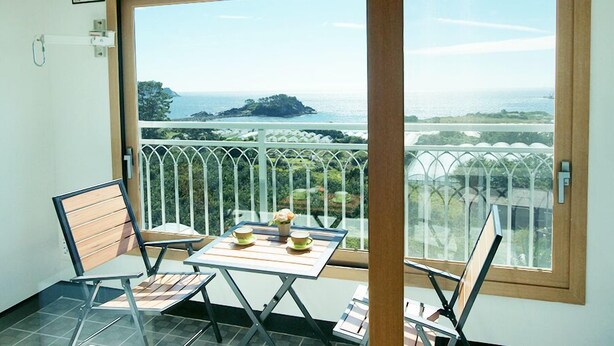 A balcony with a table and chairs, overlooking a coastal landscape with a view of the sea and a small island. - Marine Port Resort (Seogwipo)