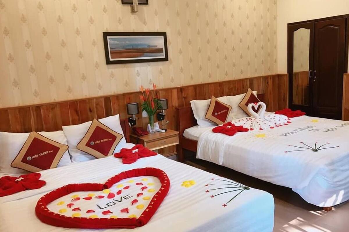 A hotel room with two beds, each with a heart-shaped decoration and a 'Love' sign. There are framed pictures on the headboard, a nightstand with a lamp, and a wooden wardrobe.