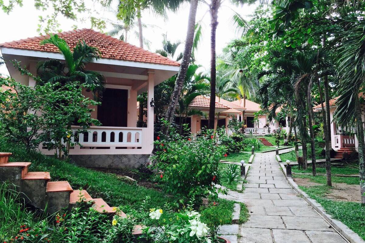 A pathway leading to a row of houses with terracotta roofs, surrounded by lush greenery and palm trees.