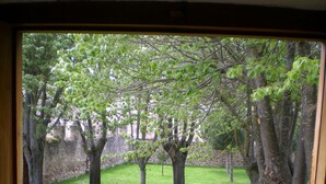 Vista a través de una ventana de un patio cubierto de césped, con árboles y un muro de piedra.