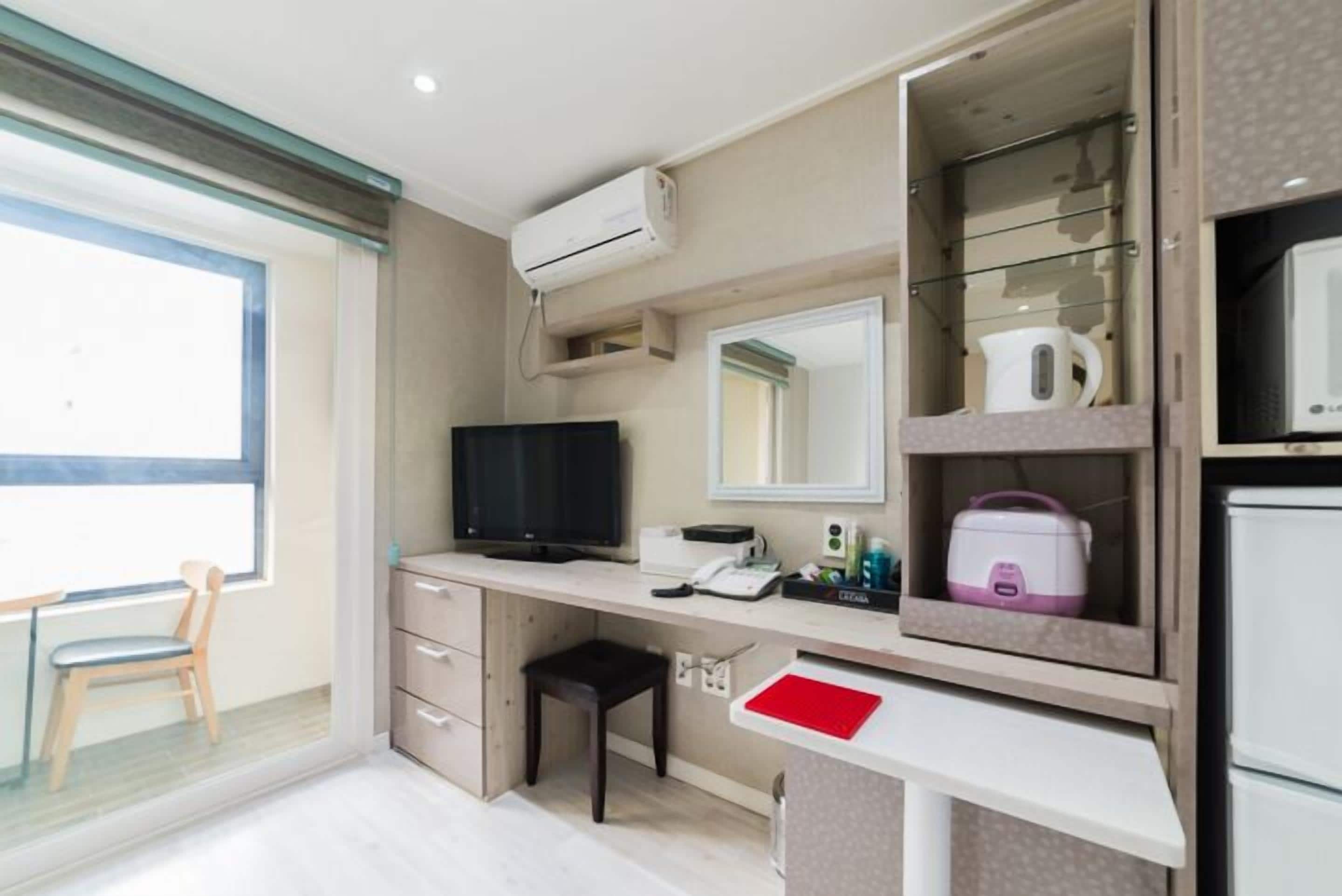 Una habitación de hotel compacta con escritorio, televisor, espejo, kitchenette y ventana.