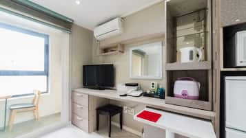 Una habitación de hotel compacta con escritorio, televisor, espejo, kitchenette y ventana.