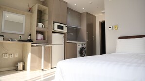 Una habitación de hotel funcional con cama, kitchenette, lavadora y un pequeño cubo de basura.