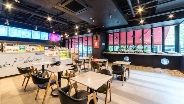 Un café moderno con una barra que exhibe diferentes opciones de comida, múltiples zonas de estar con mesas y sillas, y amplios ventanales con vistas al exterior.