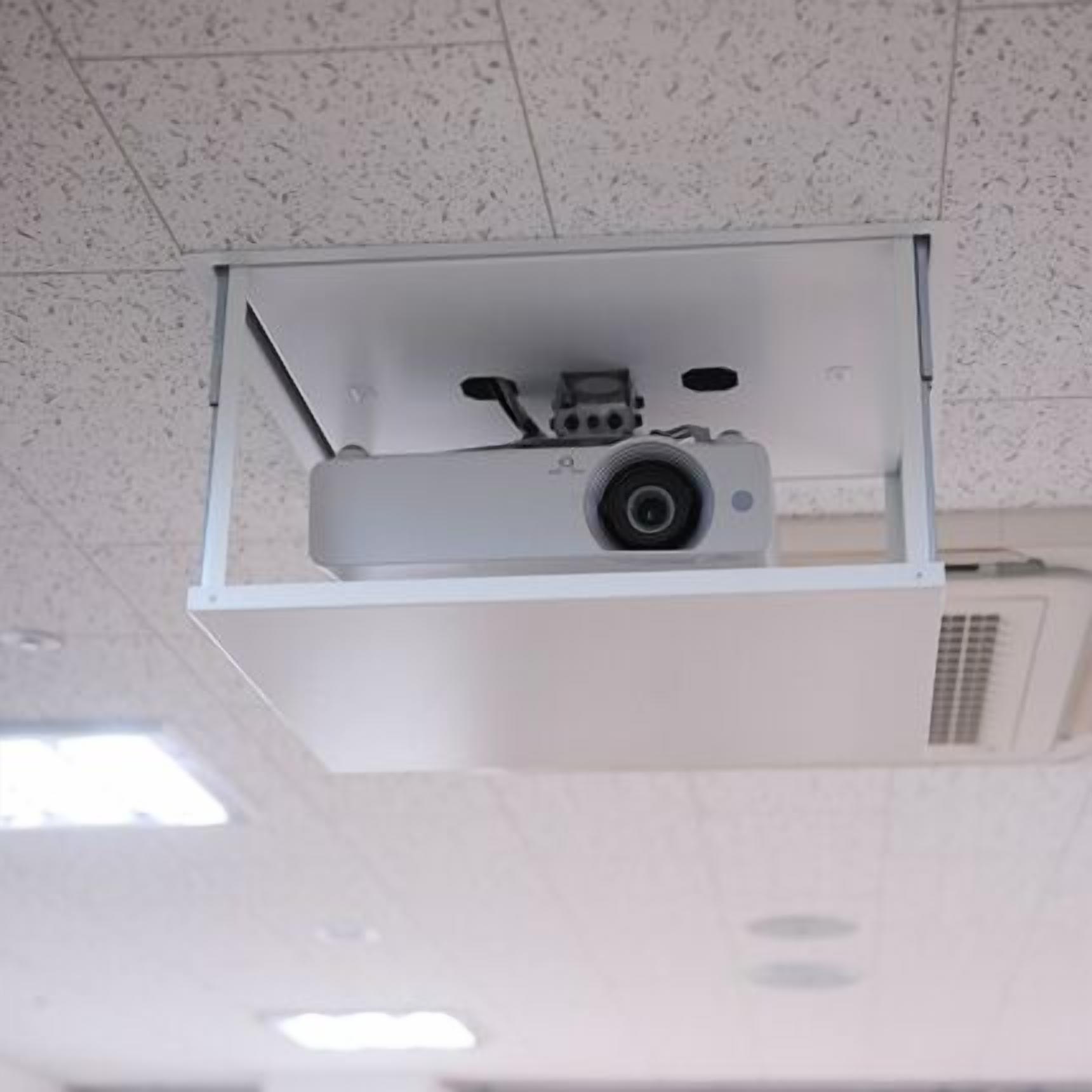 A ceiling-mounted projector in a room with a speckled ceiling and a vent.