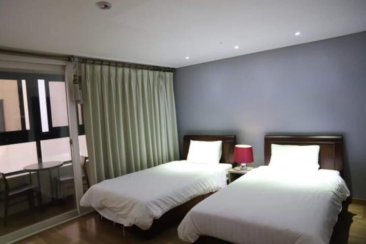 A hotel room with two beds, a window with curtains, a small table, and a nightstand with a lamp.