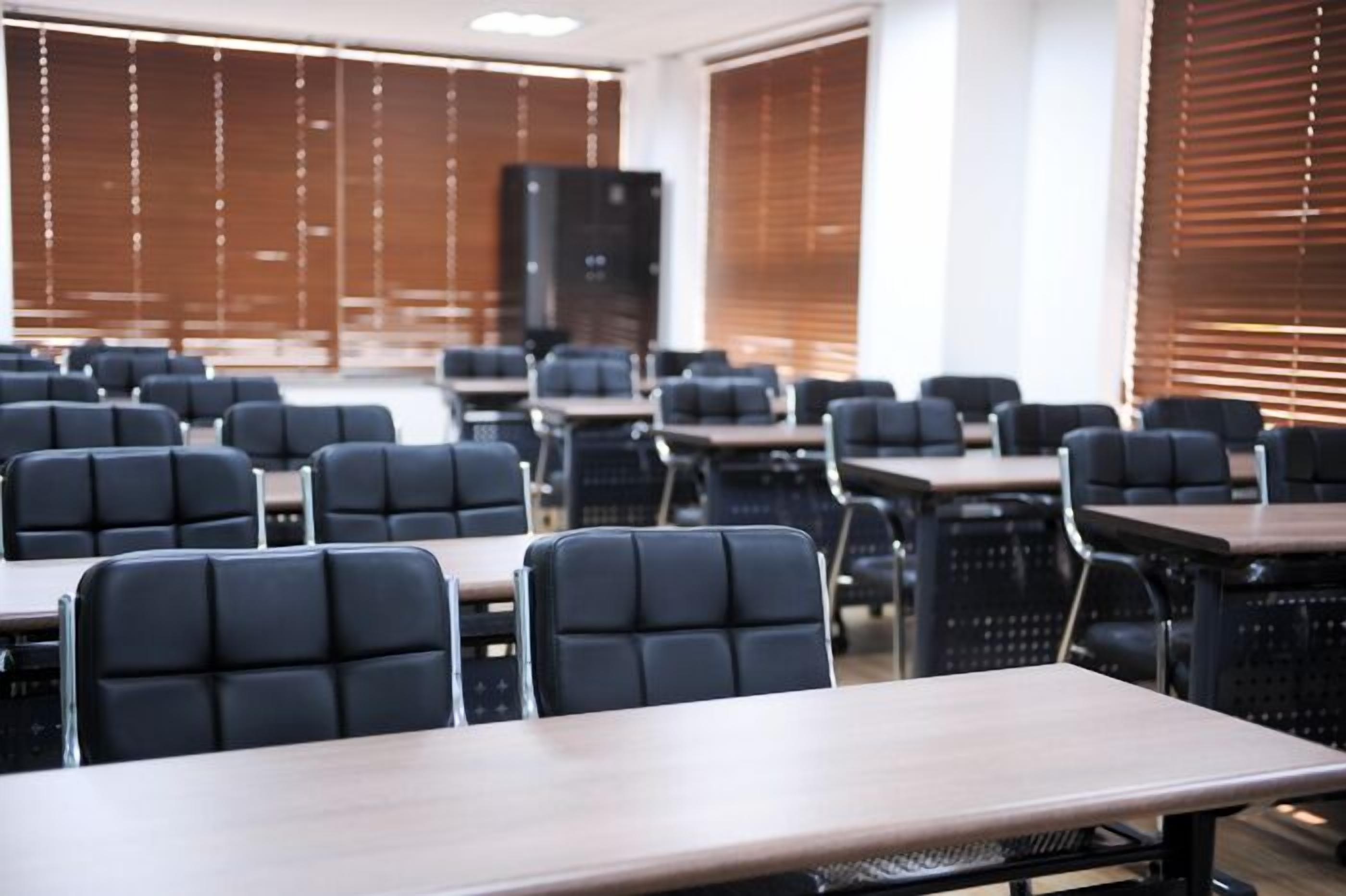 A conference room with rows of tables and chairs, large windows with blinds, and a speaker in the back.