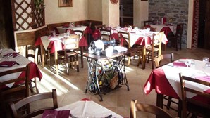 A restaurant with multiple tables and chairs, each covered with a red tablecloth. There is a vintage sewing machine in the center, surrounded by a basket of fruits and vegetables. The walls are decorated with a painting and a circular emblem. - Locanda Aquila Bianca (Valprato Soana)