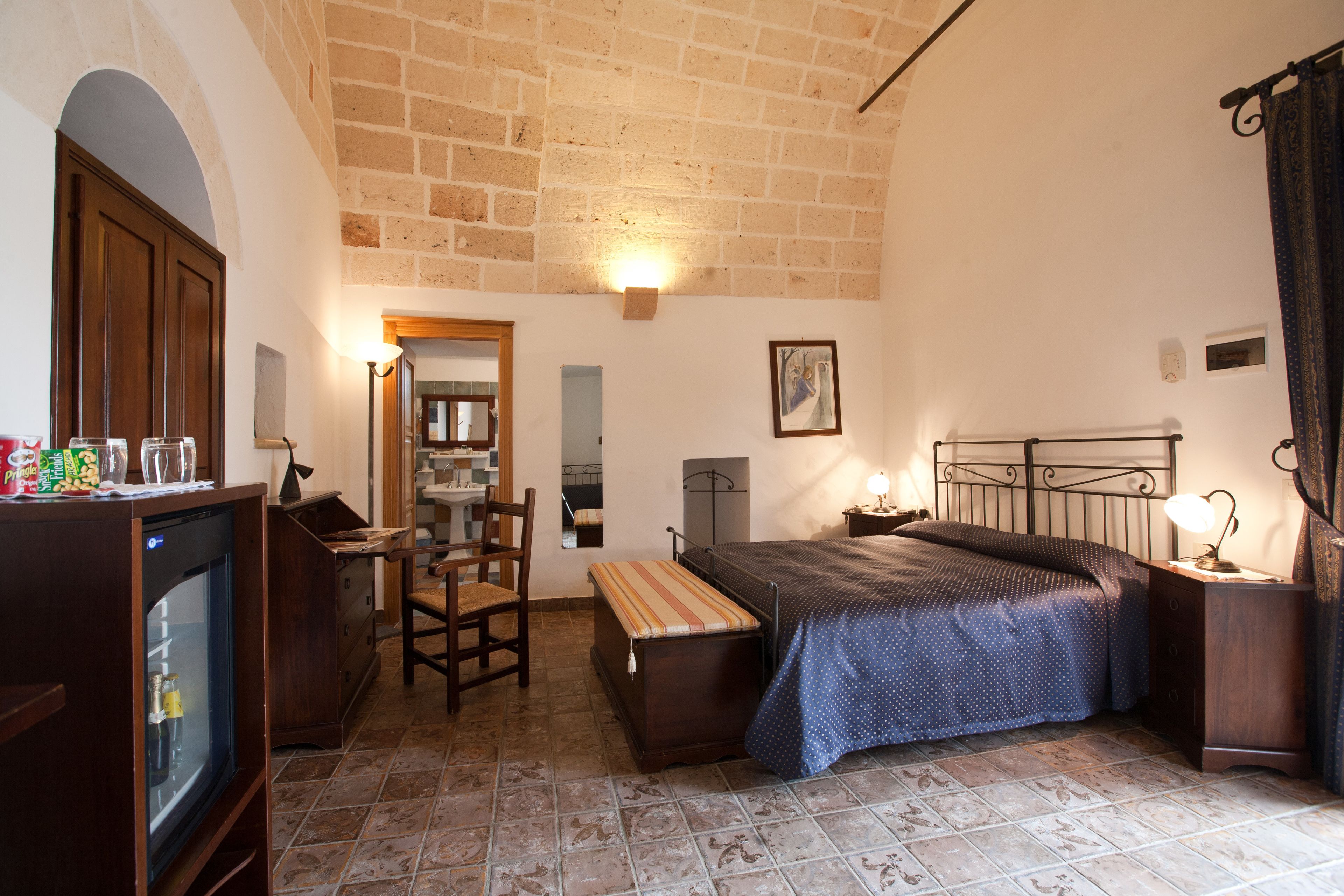 Une chambre confortable avec un lit, une table de chevet, une lampe et un petit banc. On trouve une commode en bois et un téléviseur sur le côté gauche. La pièce est dotée d’un sol carrelé et d’un plafond en briques.