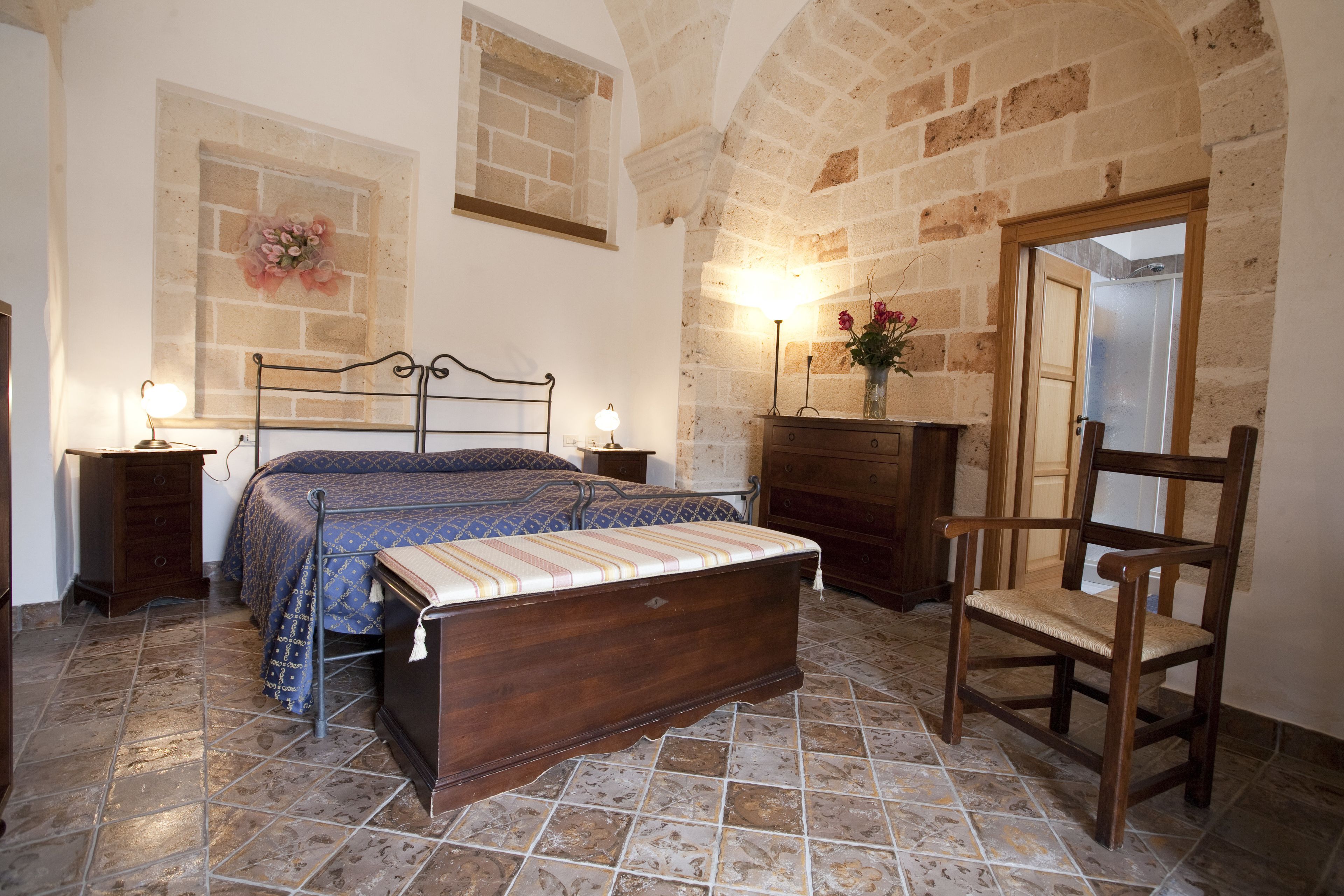 Un dormitorio con cama, mesitas de noche, una silla de madera, cómoda y pared de piedra.