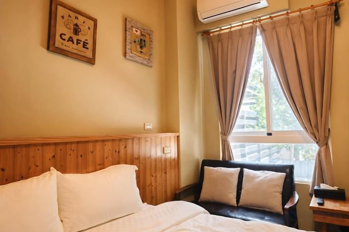 A hotel room with a bed, a wooden headboard, a window with curtains, and a wall-mounted air conditioner.