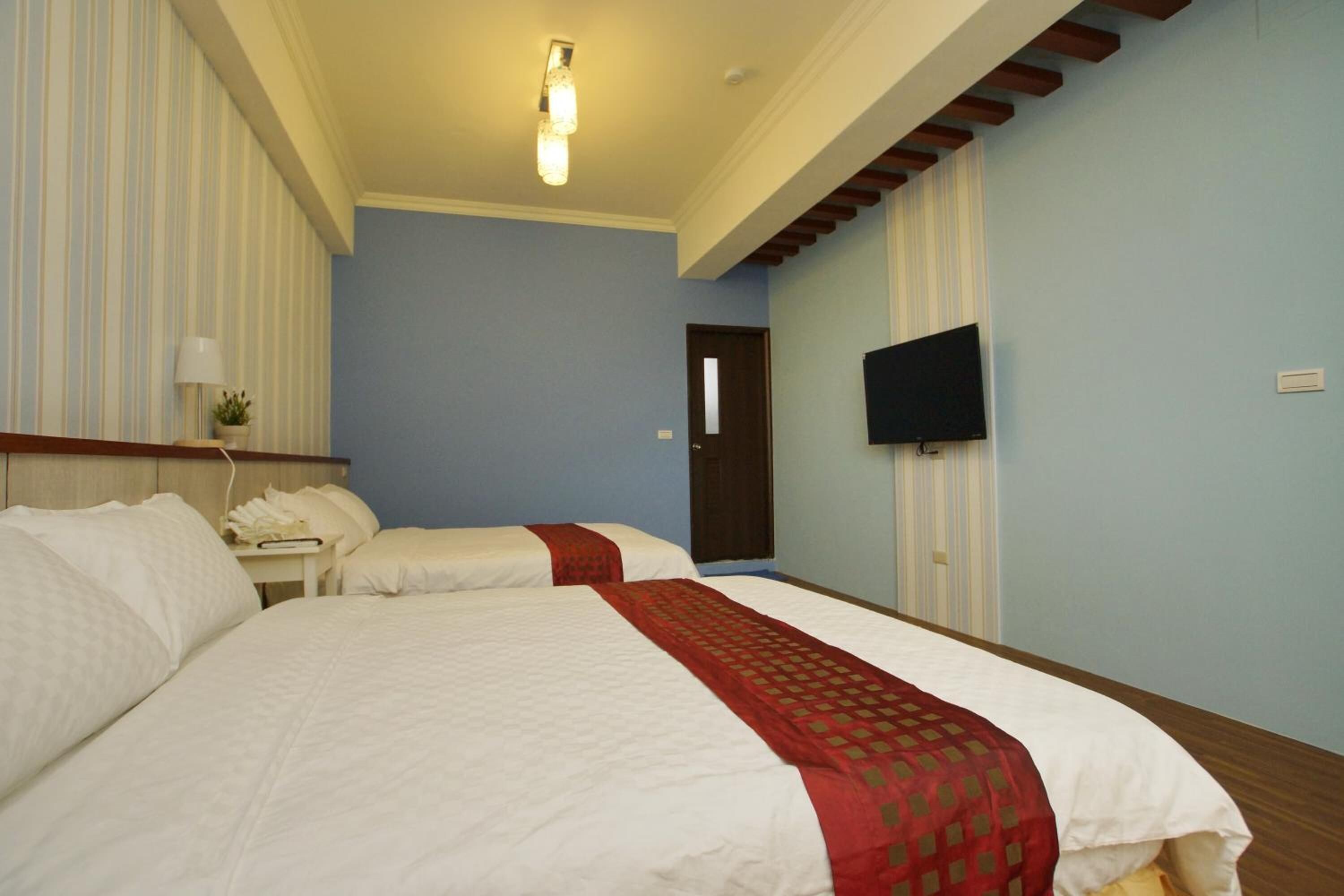 A hotel room with two beds, a TV, a bedside table with a lamp, and a window with curtains.