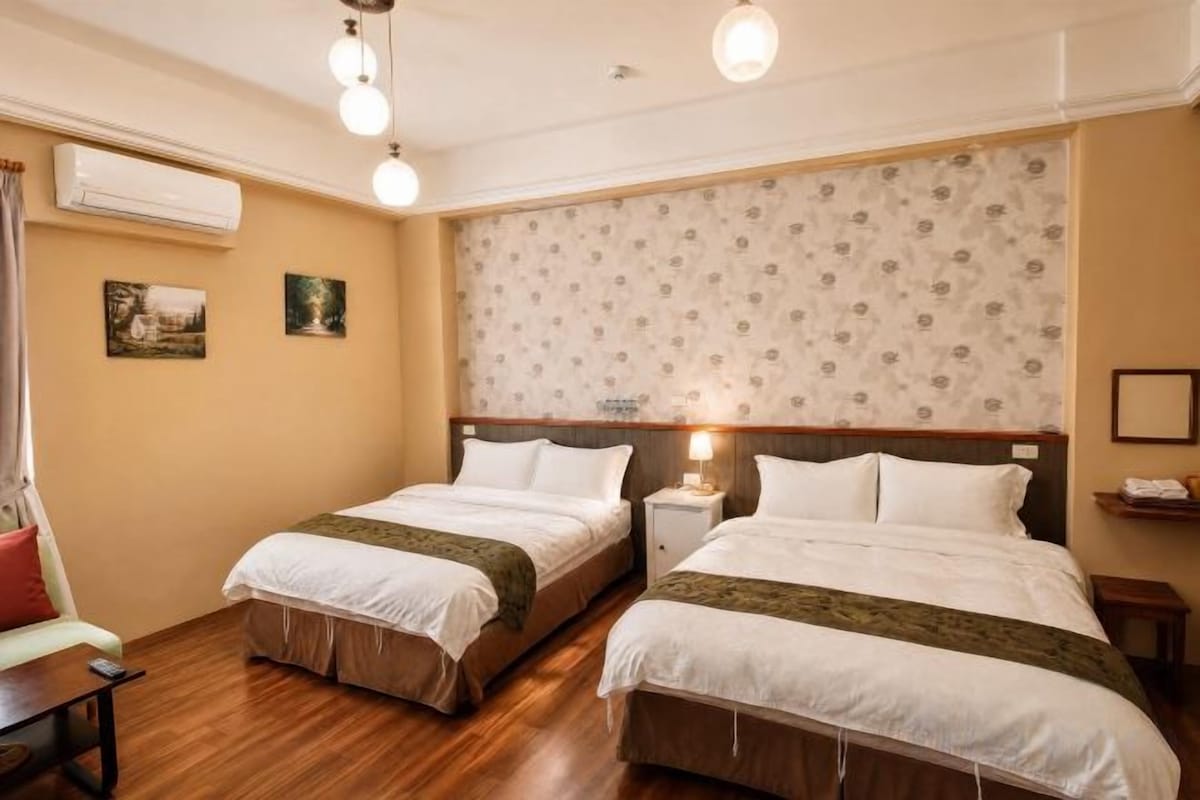 A hotel room with two beds, a bedside table with a lamp, a small desk, and a chair.
