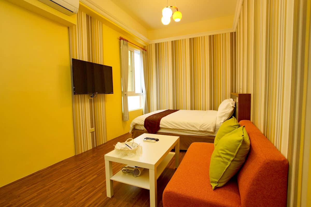 A hotel room with a bed, a sofa, a TV, and a table with a remote control and a bag.