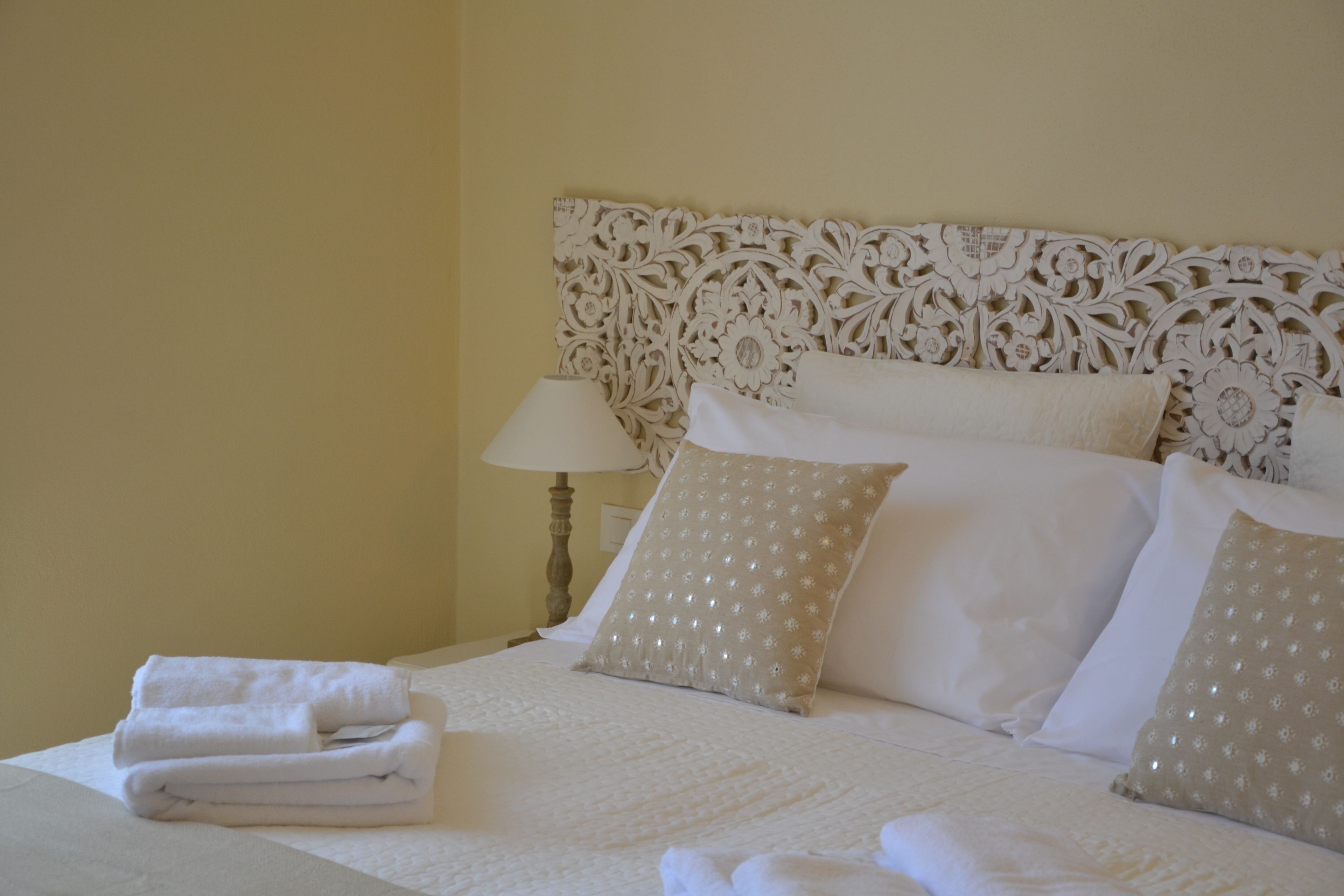 Una cama bien hecha con sábanas blancas y almohadas decorativas, una lámpara de noche y un cabecero floral en la pared.