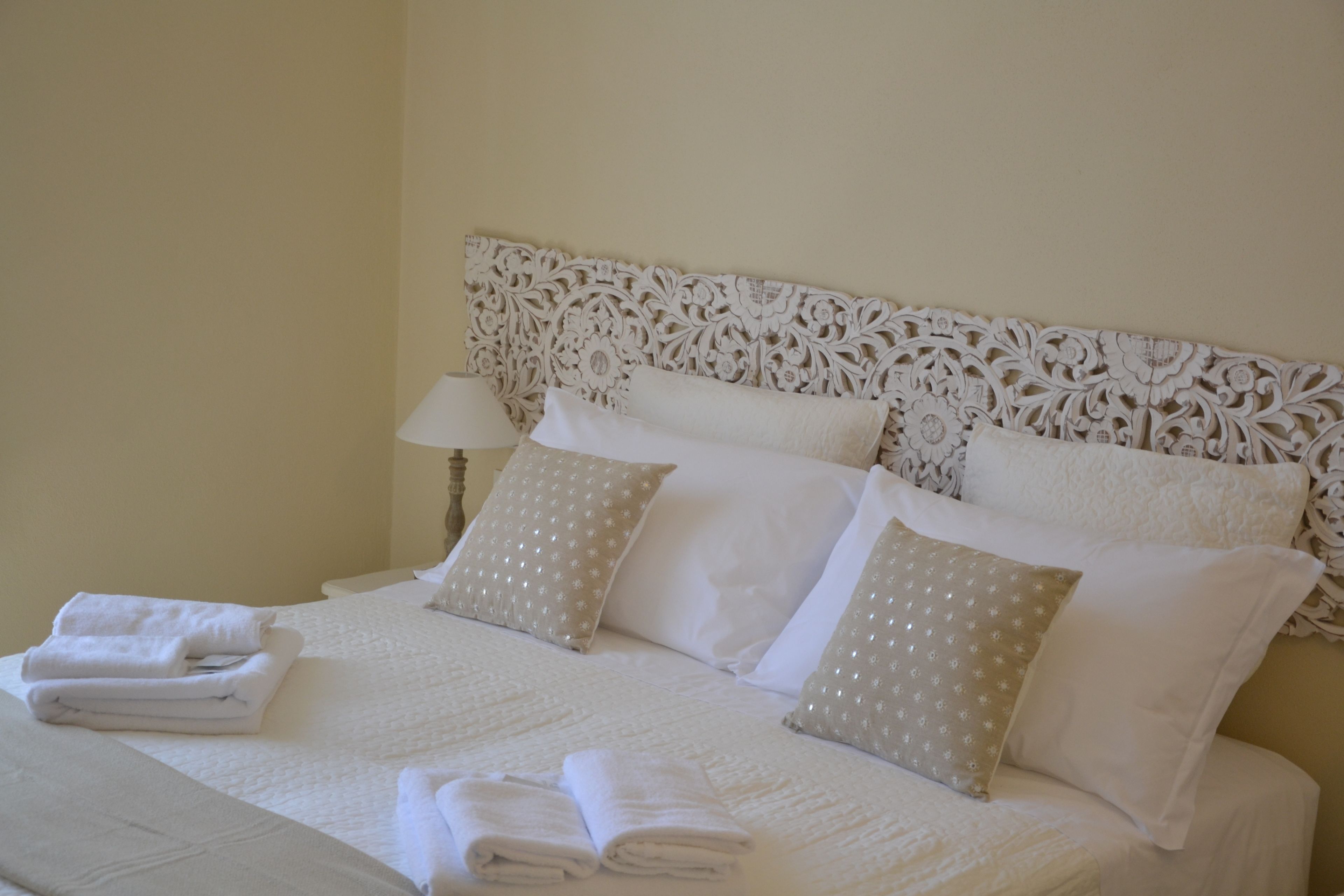A neatly made bed with white linens and decorative pillows, a headboard with intricate carvings, and a bedside table with a lamp.