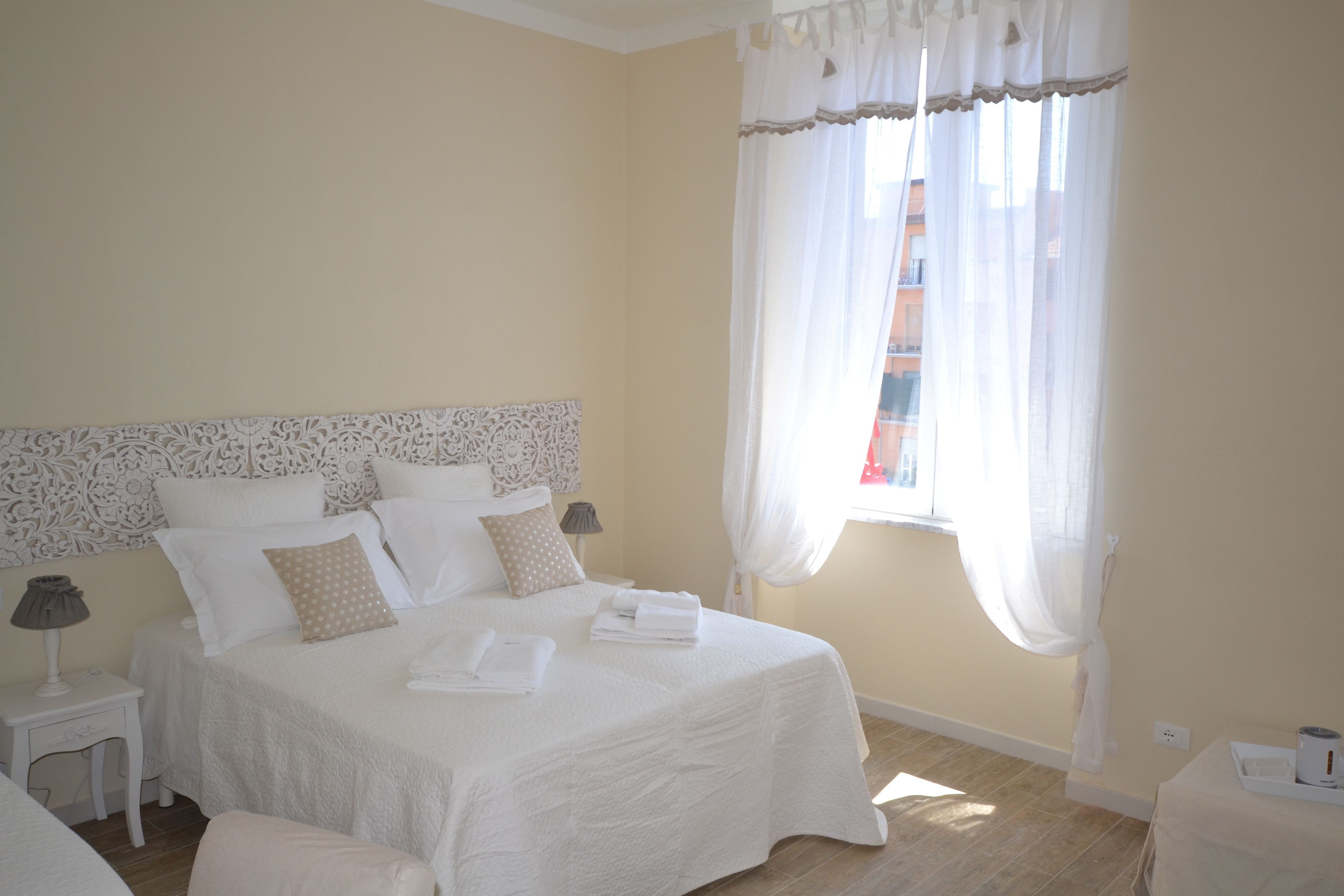 Une chambre à coucher avec un grand lit, du linge de lit blanc, une table de chevet avec une lampe et une fenêtre ornée de rideaux légers.