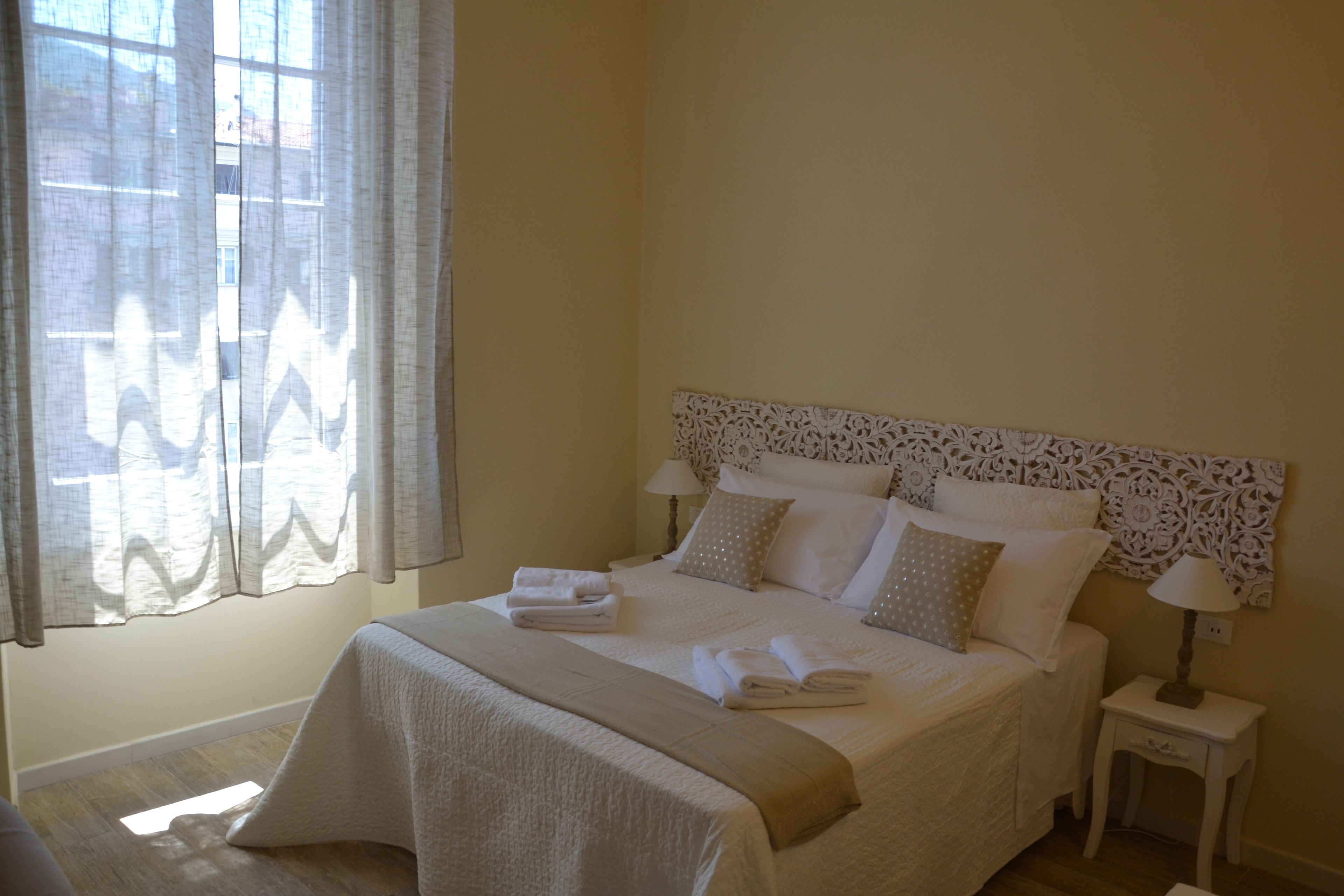 Una cama bien hecha con cabecera, una mesita de noche con lámpara y una ventana con cortinas.
