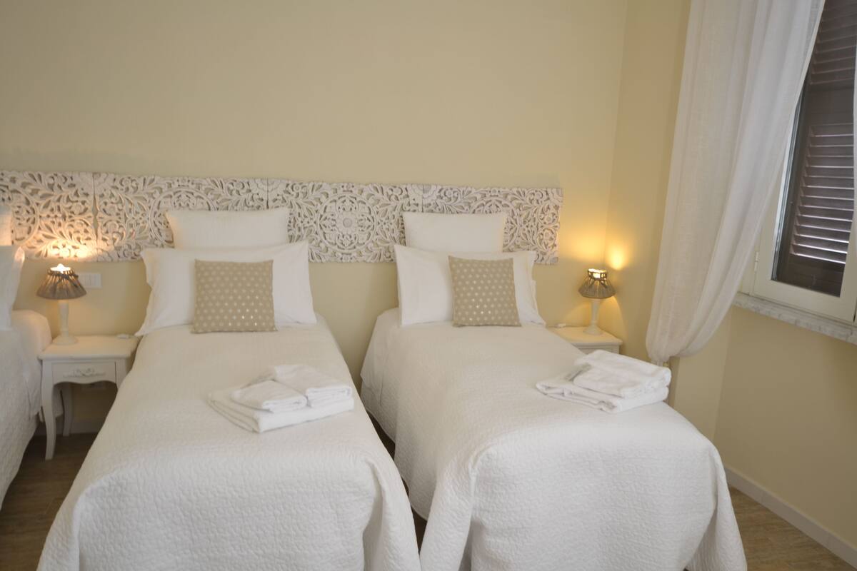 Une chambre d’hôtel avec deux lits, chacun doté de draps et d’oreillers blancs, d’une tête de lit décorative, de lampes de chevet et d’une fenêtre avec des rideaux.