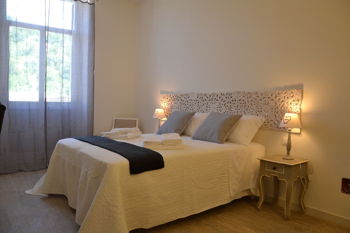 Un lit bien fait, recouvert d’une courtepointe blanche et agrémenté d’oreillers, une table de chevet avec une lampe, et une fenêtre avec des rideaux.