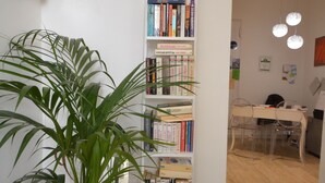 Een kamer met een witte boekenkast vol boeken, een plant in pot en een eethoek met een tafel en stoelen.