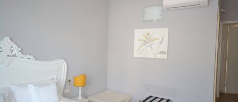 A hotel room with a bed, a bedside table with a lamp, an air conditioning unit, a towel rack, and a framed picture on the wall.