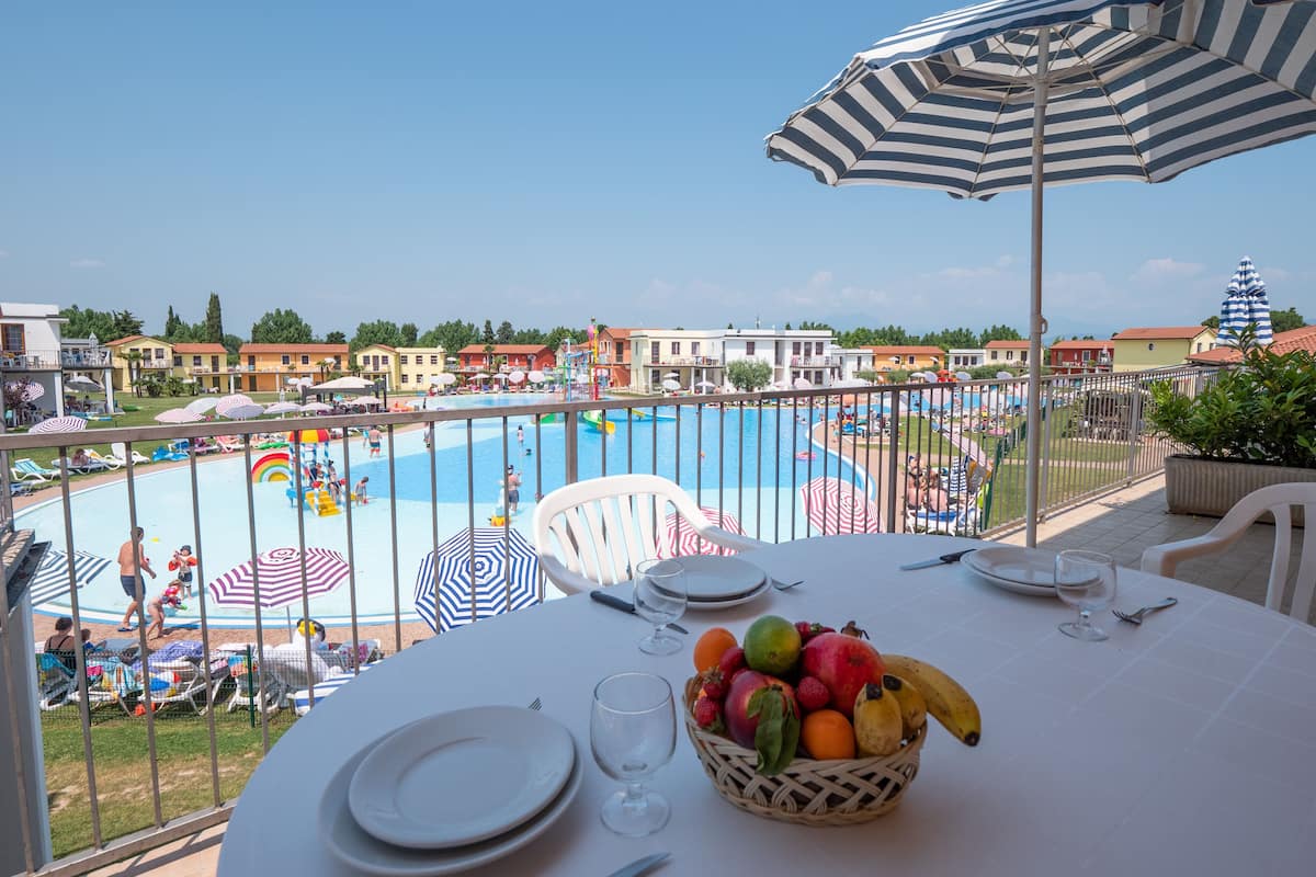 Une table à manger avec un panier de fruits, des assiettes et des couverts, donnant sur une piscine agrémentée de parasols et de chaises longues.