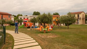 Un'area giochi per bambini con altalene e scivoli, circondata da un prato e un vialetto lastricato.