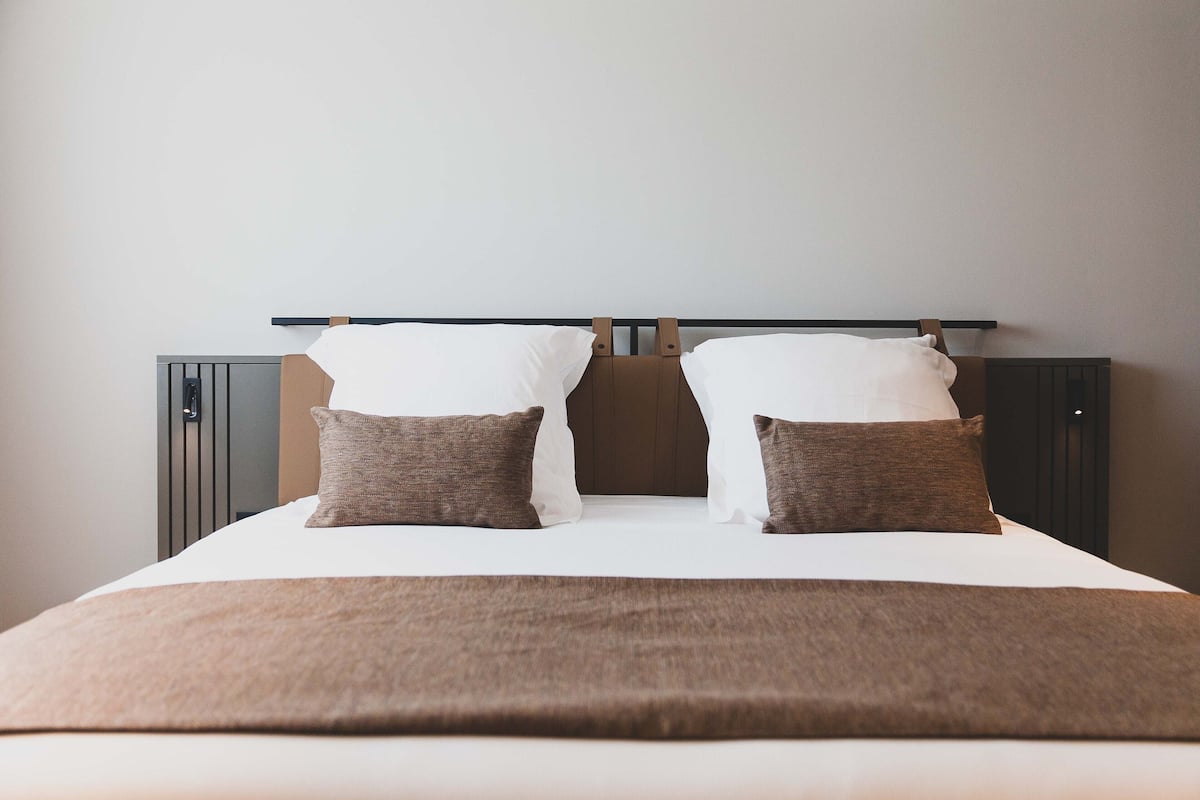 A neatly made bed with a brown blanket and two pillows, a headboard, and a wall-mounted shelf.