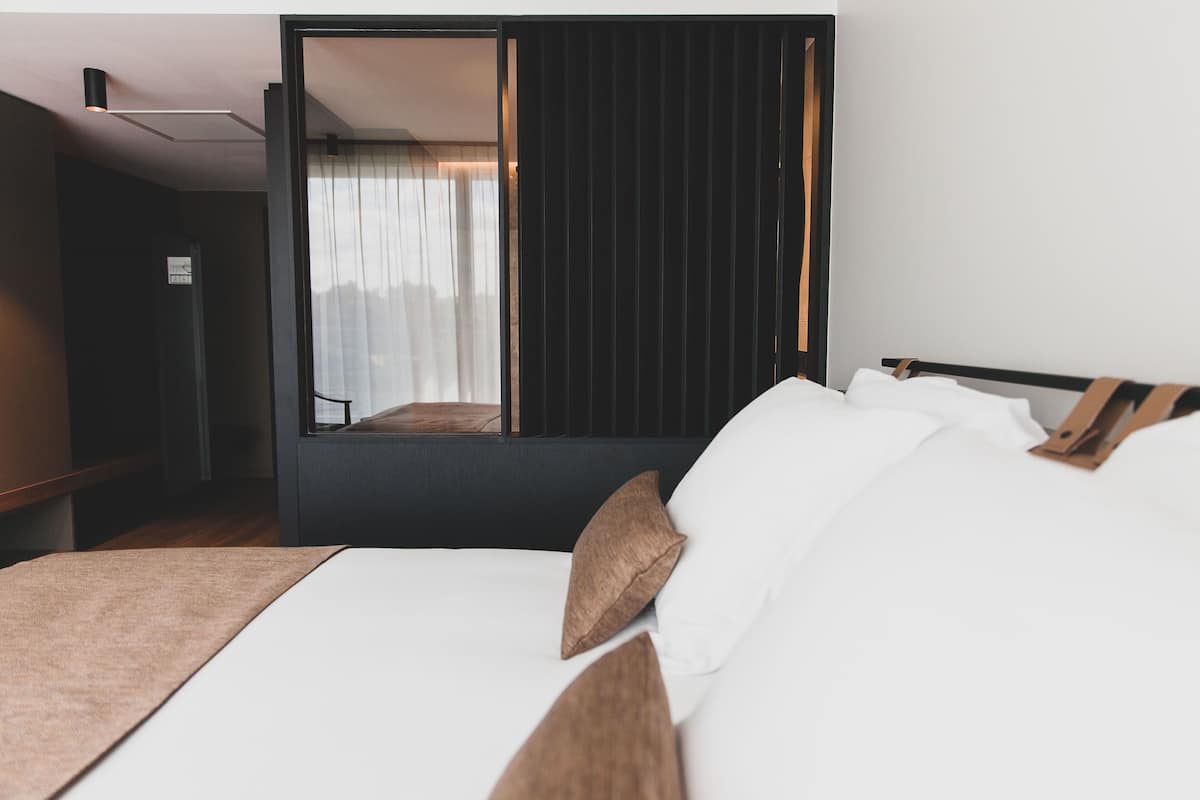 A modern hotel room with a bed, a wooden headboard, and a large window with curtains.
