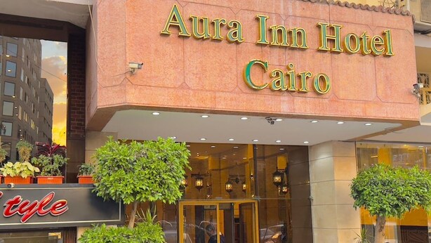 Exterior del hotel Aura Inn en El Cairo, con una entrada destacada, plantas en macetas y un letrero visible de un salón de belleza cercano.