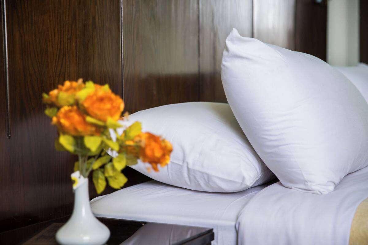 A bed with white bedding and pillows, a white vase with orange flowers, and a wooden headboard.
