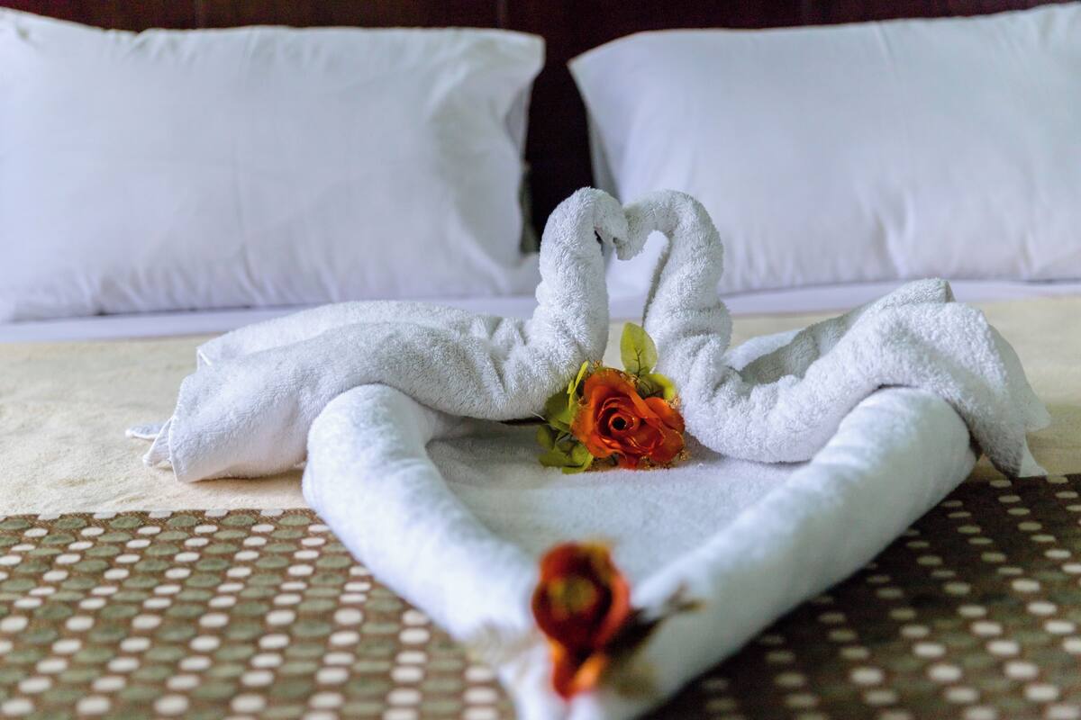 A neatly made bed with a towel arranged in the shape of a swan, a single rose, and a heart shape towel on the bed.