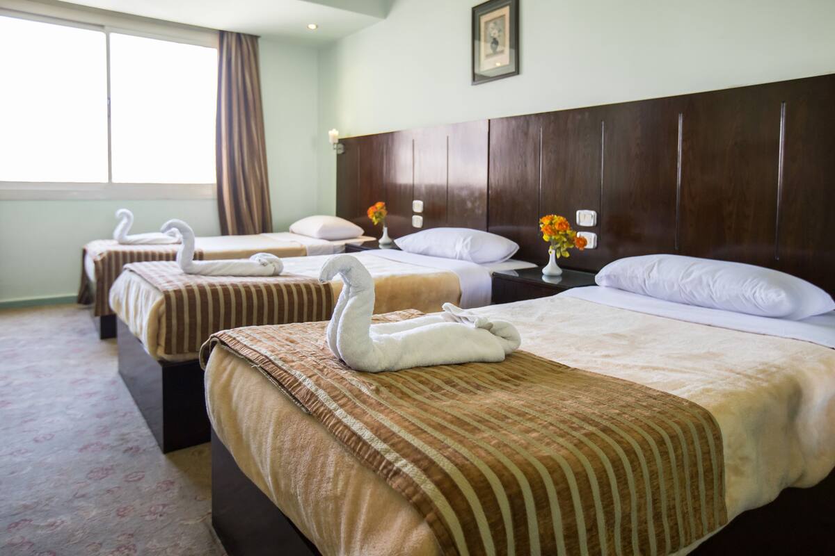 A hotel room with three beds, each with a swan-shaped towel, a large wooden headboard, and a window with curtains.