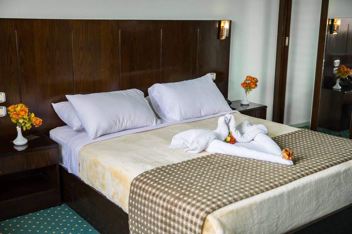 A neatly made bed with white linens, a brown checkered bedspread, and a small vase with orange flowers on the nightstand.